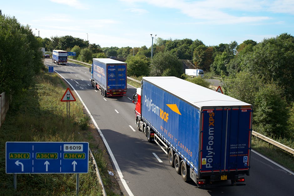 A convoy of three large freight trucks with blue and white Polyfoam DOOF trailers are moving along a multi-lane road during daylight, surrounded by green trees and under a partly cloudy sky. The trucks are positioned in the right and middle lanes with the closest truck in the foreground, showing a rear view with visible wheels, signage, and reflective strips. The road features white dashed lane markings and a blue directional road sign indicating routes A38(E), A38(W), and B6019. On the left side, there's a red triangular warning sign and a smaller rectangular informational sign. The scene captures the loading process typical of house removals and home relocation logistics, with the trucks likely transporting furniture and boxes secured within the trailers. The environment suggests a suburban or semi-rural setting, with the trucks approaching or passing through an area managed by Man With a Van Tottenham, providing professional removals and transport services in the Tottenham area.
