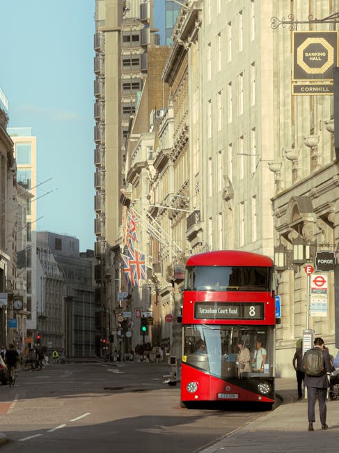 A red double-decker bus traveling along a busy city street in Tottenham, London, during daylight hours. The bus displays route number 8 and is situated near the sidewalk, where pedestrians are walking, some carrying bags and backpacks. Surrounding the bus are tall, historic-looking buildings with detailed facades, large windows, and several flags, including a Union Jack, hanging from the buildings. The street is lined with lampposts, traffic lights, and signage, with a clear blue sky overhead. This scene captures the typical urban environment in Tottenham, suitable for illustrating house removals or relocation services involving furniture transport and loading processes in busy city streets, as handled by companies like Man With a Van Tottenham.