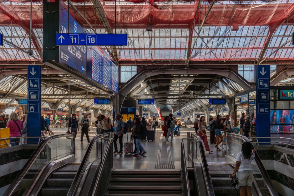 Interior view of a busy train station with a high arched glass ceiling allowing natural light to illuminate the waiting area. The platform is filled with numerous commuters, many carrying luggage, bac