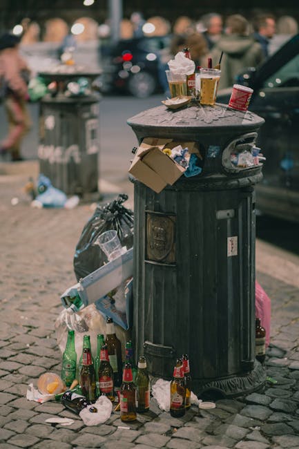 An outdoor scene showing a large pile of mixed waste, including black plastic bin liners filled with domestic rubbish, discarded packaging materials, and a yellow plastic container, all situated on a 